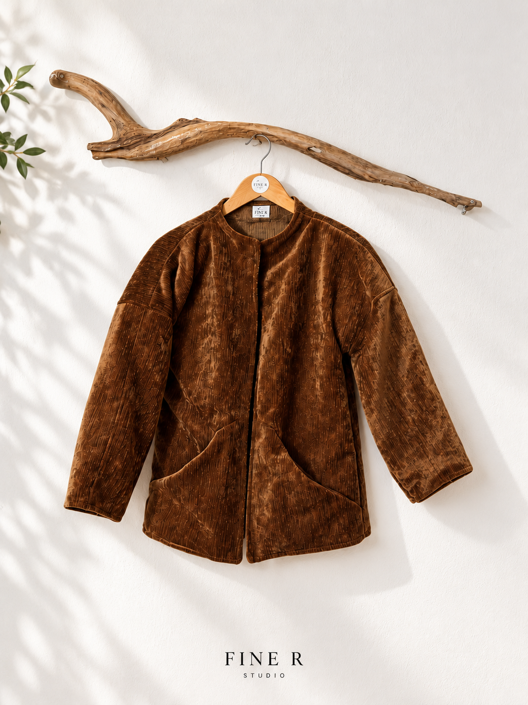 Fine R Studio copper-brown textured jacket with an open front and curved pockets, displayed on a wooden hanger beneath driftwood.