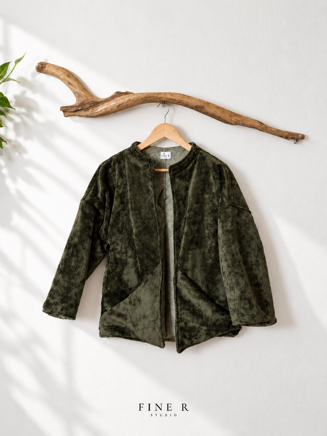 Fine R Studio olive textured open-front jacket with curved pockets, displayed on a wooden hanger beneath driftwood.